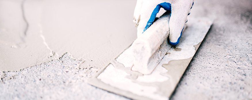 A gloved hand uses a trowel to smooth wet cement on a rough concrete surface during a concrete driveway repair.