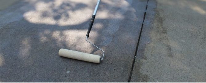 A paint roller sits on a concrete surface, partially wet, showing a clear line between the coated and uncoated areas—perfect for showcasing the results of professional concrete driveway repair. Shadows from trees are visible in the background.