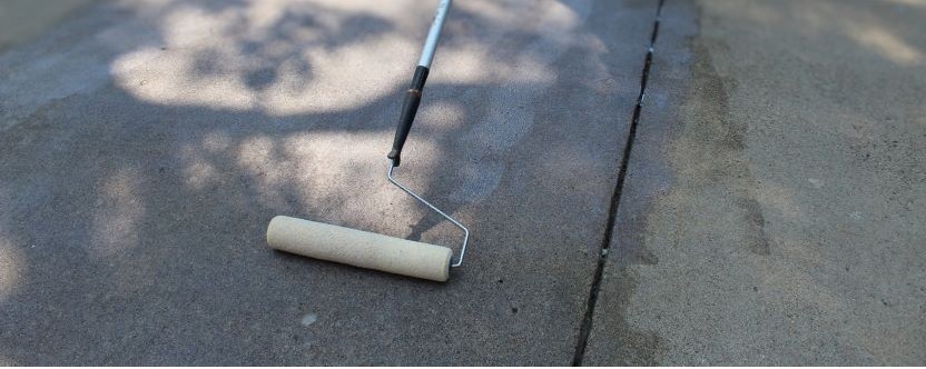 A paint roller sits on a concrete surface, partially wet, showing a clear line between the coated and uncoated areas—perfect for showcasing the results of professional concrete driveway repair. Shadows from trees are visible in the background.