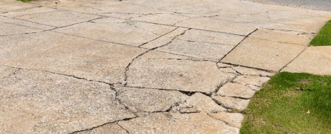 Cracked concrete driveway with multiple deep fissures and uneven surfaces, bordered by patches of green grass—ideal for a residential concrete company specializing in concrete driveway repair.