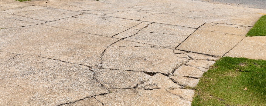 Cracked concrete driveway with multiple deep fissures and uneven surfaces, bordered by patches of green grass—ideal for a residential concrete company specializing in concrete driveway repair.