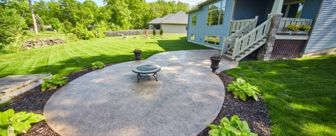 A backyard patio features a round, stamped concrete surface crafted by expert concrete service, a central fire pit, potted plants, lush grass, garden beds beyond, and a blue house with a porch to the right.