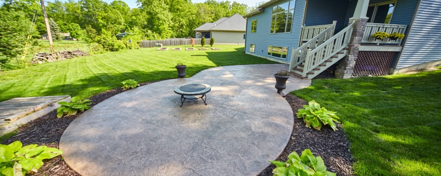A backyard patio features a round, stamped concrete surface crafted by expert concrete service, a central fire pit, potted plants, lush grass, garden beds beyond, and a blue house with a porch to the right.