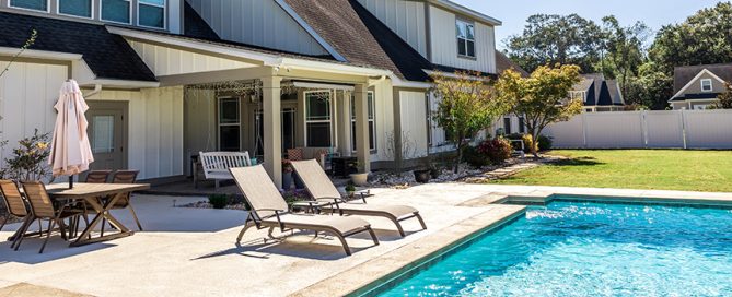 A modern backyard with a swimming pool, two lounge chairs, a dining table with an umbrella, and a porch swing—expertly crafted by a leading residential concrete company—stands before a large house with white siding and expansive windows.