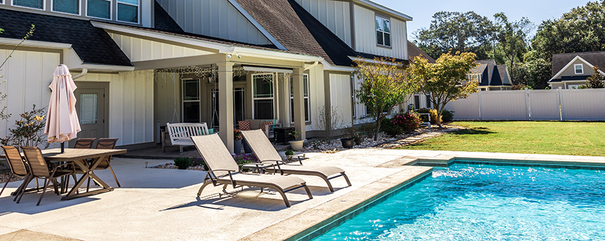The,Side,View,Of,A,Long,Craftsman,Cottage,White,Cream A modern backyard with a swimming pool, two lounge chairs, a dining table with an umbrella, and a porch swing—expertly crafted by a leading residential concrete company—stands before a large house with white siding and expansive windows.
