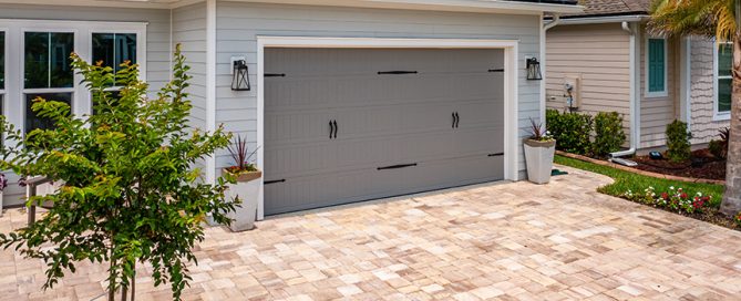 A modern house with a light gray exterior and a two-car garage featuring a gray door, black handles, and decorative lanterns. A residential concrete company paved the tan brick driveway, with potted plants and landscaping enhancing the entrance.