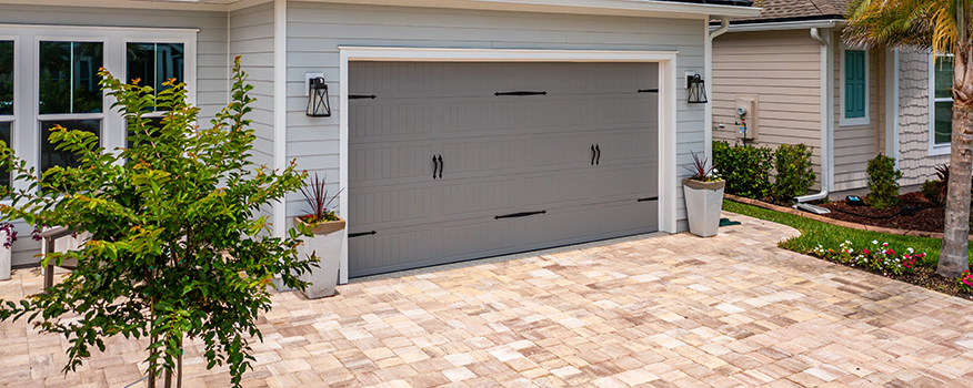 A modern house with a light gray exterior and a two-car garage featuring a gray door, black handles, and decorative lanterns. A residential concrete company paved the tan brick driveway, with potted plants and landscaping enhancing the entrance.