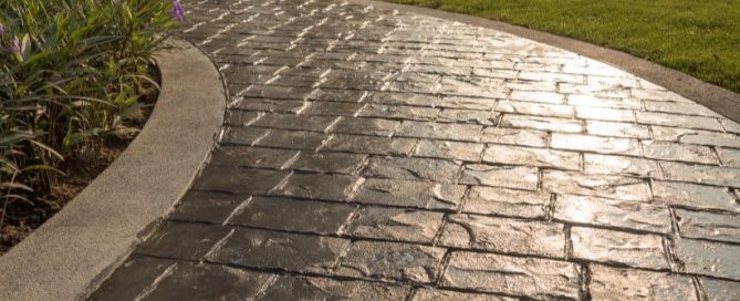 Curved stone pathway bordered by concrete edges, showcasing expert concrete service. Green grass lines one side, while purple flowers brighten the other, all illuminated by sunlight.