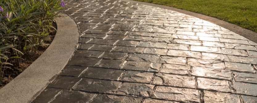 stamped-concrete-st-petersburg Curved stone pathway bordered by concrete edges, showcasing expert concrete service. Green grass lines one side, while purple flowers brighten the other, all illuminated by sunlight.