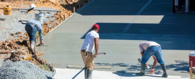 Three construction workers from a residential concrete company smooth and finish wet concrete on a large driveway during the day, surrounded by construction materials and gravel.