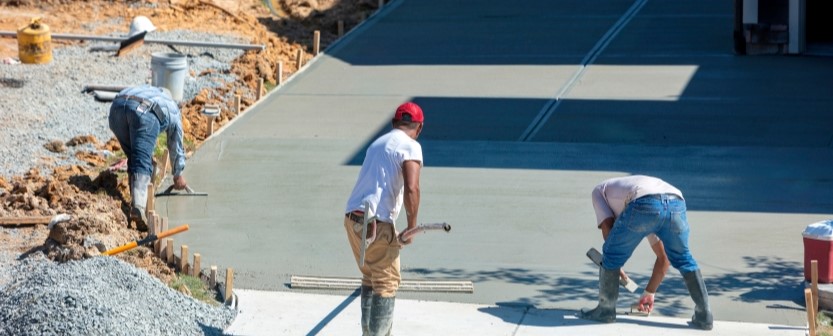 Three construction workers from a residential concrete company smooth and finish wet concrete on a large driveway during the day, surrounded by construction materials and gravel.
