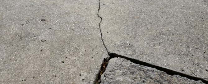 A close-up view of a cracked concrete surface reveals a jagged line running through the middle—a common sight before concrete driveway repair or other professional concrete service is performed. A broken piece sits at the bottom right corner.
