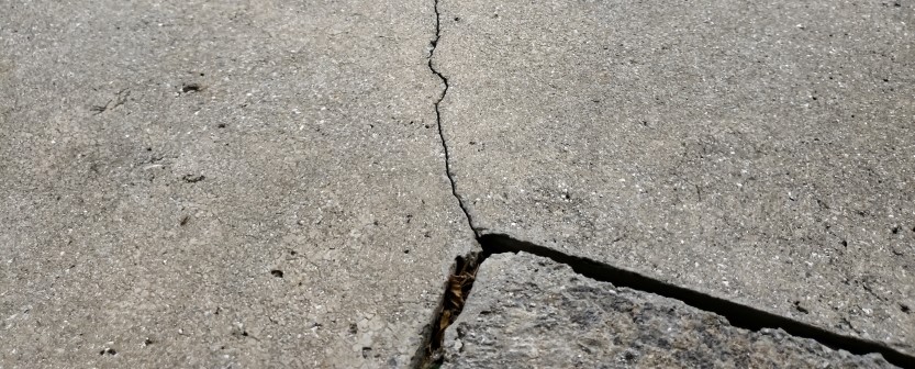 A close-up view of a cracked concrete surface reveals a jagged line running through the middle—a common sight before concrete driveway repair or other professional concrete service is performed. A broken piece sits at the bottom right corner.