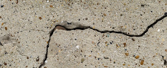 A close-up view of a cracked concrete surface, captured by a residential concrete company, shows a jagged black crack running through the light-colored, rough texture with small pebbles embedded in it.
