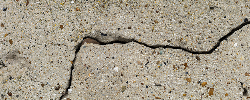 A close-up view of a cracked concrete surface, captured by a residential concrete company, shows a jagged black crack running through the light-colored, rough texture with small pebbles embedded in it.