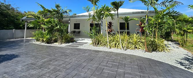 A modern white house with a metal roof is surrounded by lush tropical plants and trees, featuring a wide, clean driveway crafted by a leading residential concrete company under a clear blue sky.