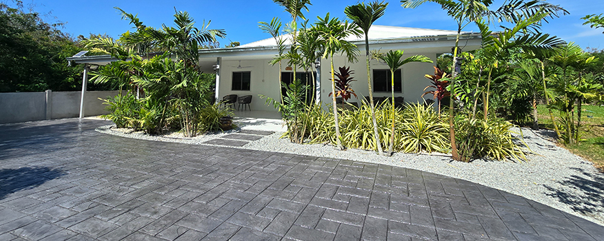 A modern white house with a metal roof is surrounded by lush tropical plants and trees, featuring a wide, clean driveway crafted by a leading residential concrete company under a clear blue sky.