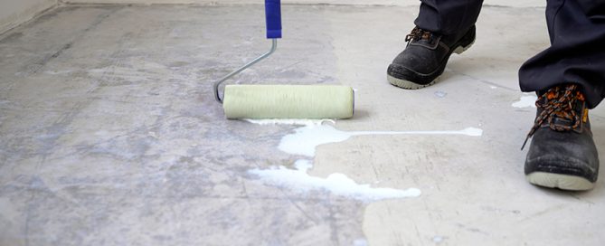 A person wearing black work boots uses a paint roller to apply a white liquid primer or sealant to a concrete floor, preparing the surface for a residential concrete company’s repair or finishing work.