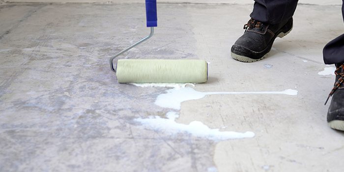A person wearing black work boots uses a paint roller to apply a white liquid primer or sealant to a concrete floor, preparing the surface for a residential concrete company’s repair or finishing work.