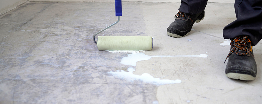 A person wearing black work boots uses a paint roller to apply a white liquid primer or sealant to a concrete floor, preparing the surface for a residential concrete company’s repair or finishing work.