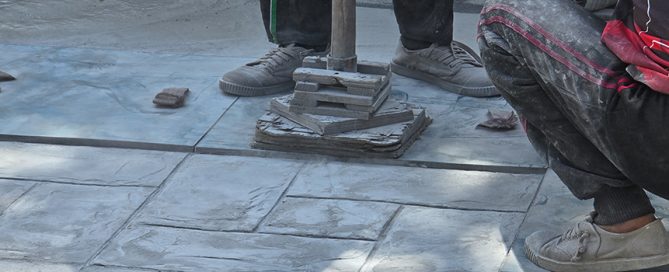 Two people wearing sneakers work on a tiled pavement. One is crouched, and the other stands beside a metal tool placed on stacked tiles—a typical scene for a concrete company Tampa relies on. The ground and their clothes are covered in dust.