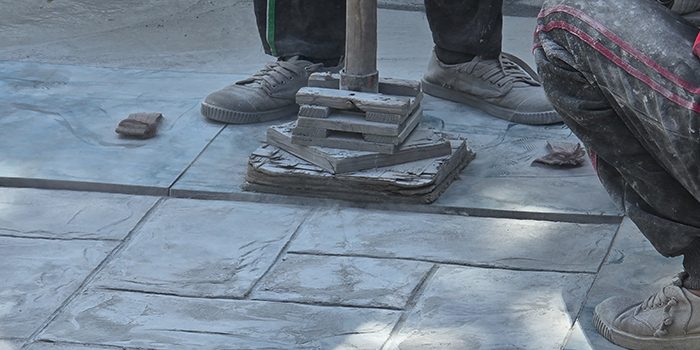 Two people wearing sneakers work on a tiled pavement. One is crouched, and the other stands beside a metal tool placed on stacked tiles—a typical scene for a concrete company Tampa relies on. The ground and their clothes are covered in dust.