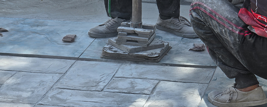 Two people wearing sneakers work on a tiled pavement. One is crouched, and the other stands beside a metal tool placed on stacked tiles—a typical scene for a concrete company Tampa relies on. The ground and their clothes are covered in dust.