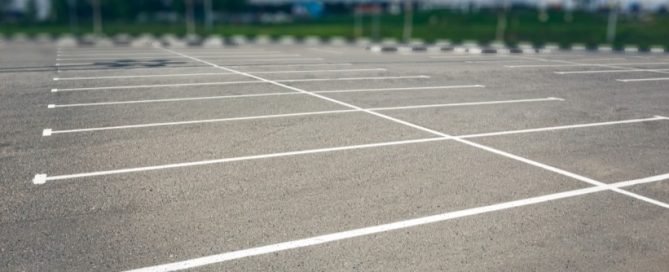 An empty parking lot with clearly marked white parking lines, freshly maintained through concrete driveway repair, no cars present, and a blurred background of greenery and distant buildings.