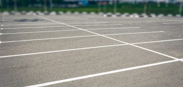 An empty parking lot with clearly marked white parking lines, freshly maintained through concrete driveway repair, no cars present, and a blurred background of greenery and distant buildings.