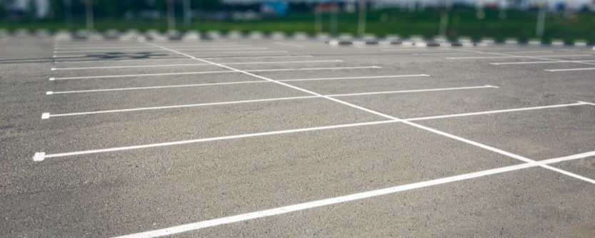 commercial-parking-lot-sidewalk-concrete-solutions An empty parking lot with clearly marked white parking lines, freshly maintained through concrete driveway repair, no cars present, and a blurred background of greenery and distant buildings.