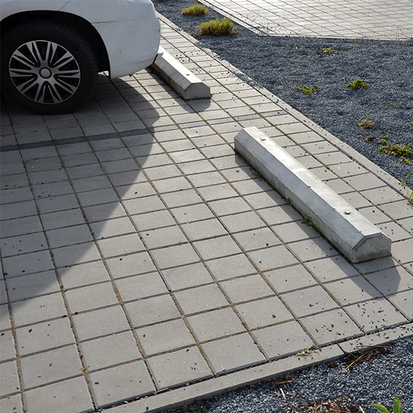 Commercial-Concrete-1 A white car is partially visible, parked next to a concrete parking stop on a paved parking lot with gray bricks—an area that could benefit from expert residential concrete company services. Small patches of grass are visible nearby.