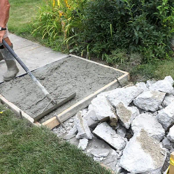 Concrete-Replacement A person spreads wet concrete with a rake on a prepared sidewalk area bordered by wood, while broken old concrete pieces pile nearby—showcasing the skilled work of a residential concrete company with lush green plants in the background.