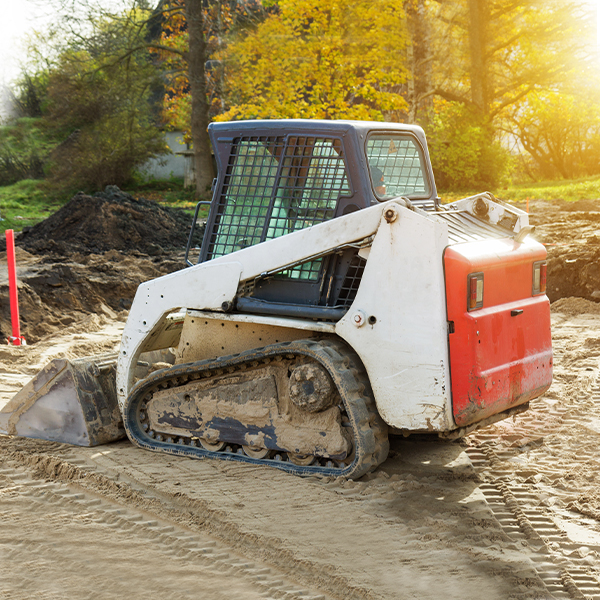 Excavation-2 A small, white and red skid-steer loader with rubber tracks is parked on sandy soil at a construction site, where a residential concrete company prepares for concrete driveway repair, surrounded by trees and sunlight in the background.