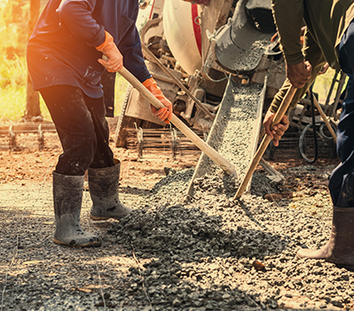 Workers in boots use shovels to spread wet concrete being poured from a cement mixer onto the ground, showcasing the expertise of a residential concrete company at work on a construction site.