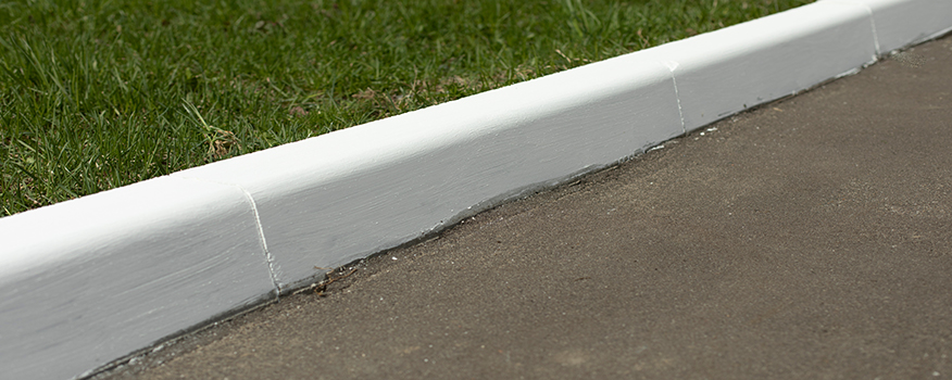 A close-up view of a freshly painted white curb, expertly crafted by a residential concrete company, separates a grassy area from a paved surface.