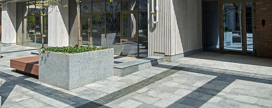 A modern outdoor area with stone tiles, a raised planter box featuring a tree, wooden benches, and large glass doors leading into a building; sunlight creates shadows on the pavement crafted by a top residential concrete company.