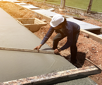 A worker in a white hard hat smooths wet concrete with a long tool at an outdoor construction site, showcasing the expertise of a residential concrete company surrounded by dirt and construction materials.