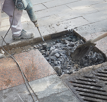 A worker from a residential concrete company uses a jackhammer to break up pavement while another person clears debris near a sidewalk drain, preparing the area for concrete driveway repair.