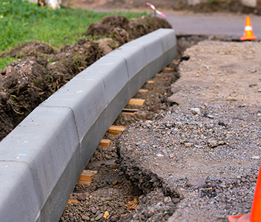 A close-up of newly installed concrete curbstones along a road under construction, completed by a professional concrete company Tampa. Wooden spacers, gravel, and orange traffic cones are visible in the background.