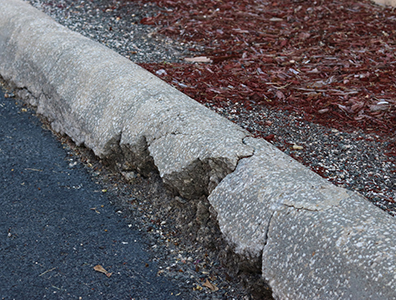A cracked and crumbling concrete curb separates an asphalt road from a landscaped area with red mulch and rocks, highlighting the need for expert concrete driveway repair by a reliable concrete company Tampa trusts.