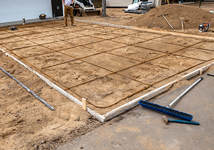 A rectangular area is prepared for concrete pouring by a concrete company in Tampa, with a grid of rebar on packed dirt and wooden forms. Construction tools and a person in work clothes are visible in the background.