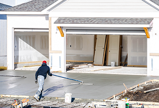 A worker in a red hat smooths freshly poured concrete on a driveway in front of a garage, showcasing expert concrete driveway repair by a residential concrete company. White doors and construction materials are visible inside the open garage.