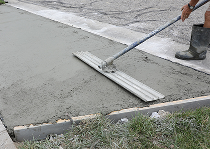A person wearing boots smooths wet concrete on a sidewalk with a long-handled tool, next to a curb and some grass—typical work for a residential concrete company.