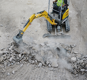A person operates a small yellow excavator breaking up concrete and rocks, with dust rising from the debris on a construction site during a concrete driveway repair.