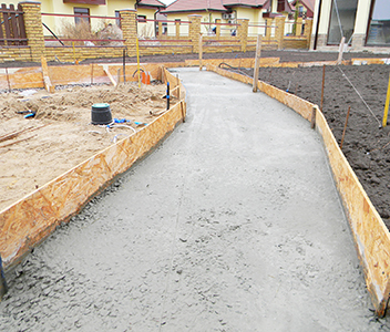 A newly poured concrete sidewalk in a residential area, bordered by wooden forms on both sides, showcases the quality work of a residential concrete company, with soil and construction materials visible nearby.