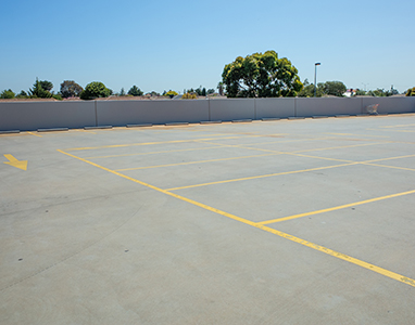 An empty outdoor parking lot with yellow lines marking spaces and a yellow arrow on the pavement, maintained by a leading residential concrete company. A low wall and trees are visible in the background under a clear blue sky.