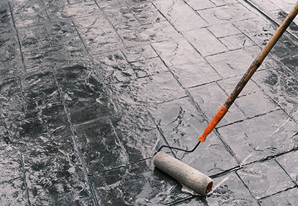 A paint roller attached to a long handle is being used to apply a coat of sealant on dark, textured, stamped concrete tiles by a residential concrete company.