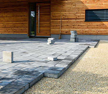 Several stacks of gray paving stones are arranged on a partially finished patio in front of a modern wooden house, where a broom leans against the wall and a gravel area lines the paved surface, prepared by a residential concrete company.