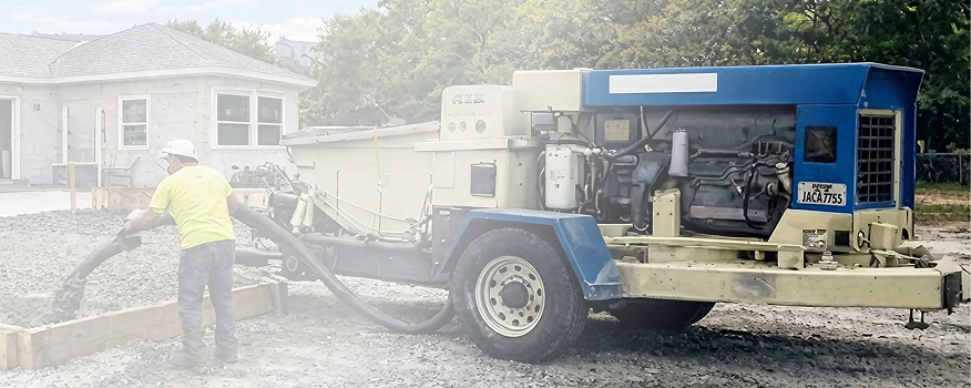 A construction worker from a residential concrete company operates a concrete pump truck, pouring concrete into a wooden frame at a building site next to a house under construction.