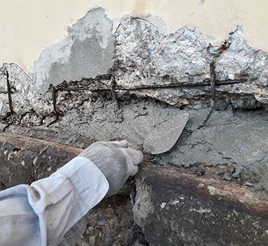 A person wearing a work glove applies fresh cement to a damaged concrete wall, covering exposed metal reinforcements and cracks with a trowel—a skilled touch often provided by a professional residential concrete company.